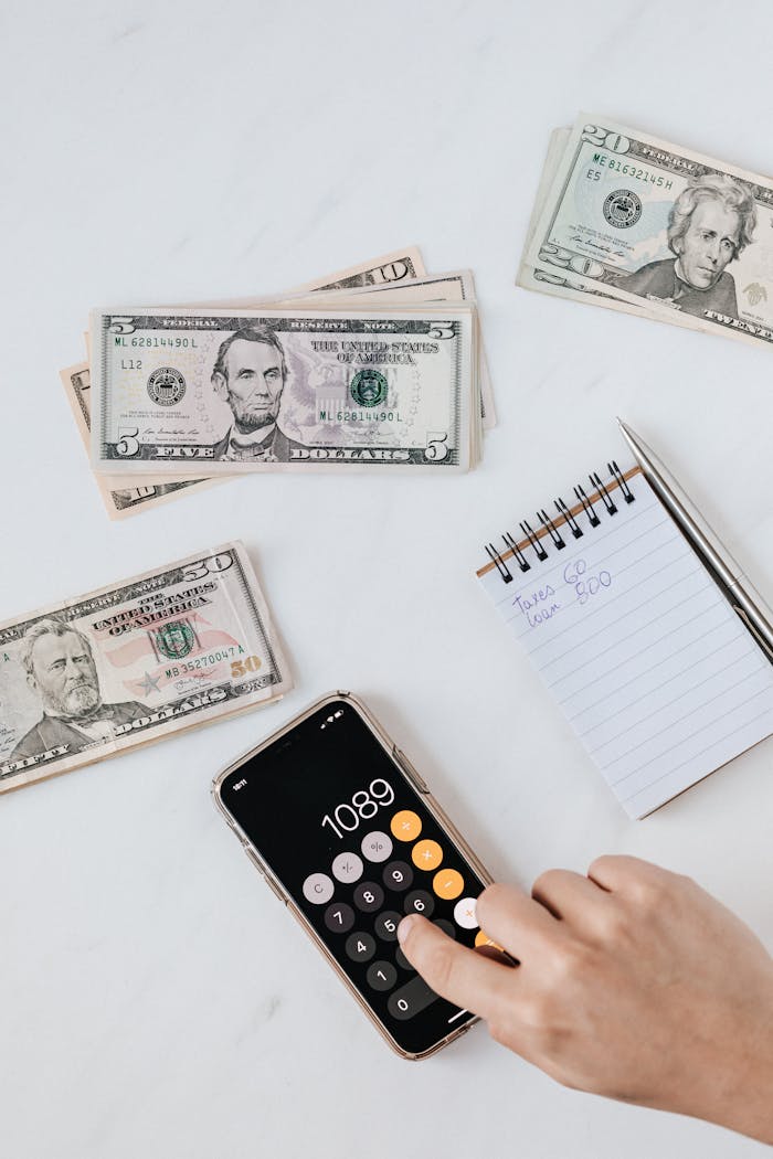 From above of crop unrecognizable person using calculator app on cellphone while calculating total amount of money sitting at table with different piles of American dollars and notebook with notes