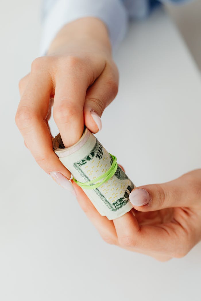 Hands holding a rolled bundle of US dollar bills bound by a rubber band, in a bright setting.